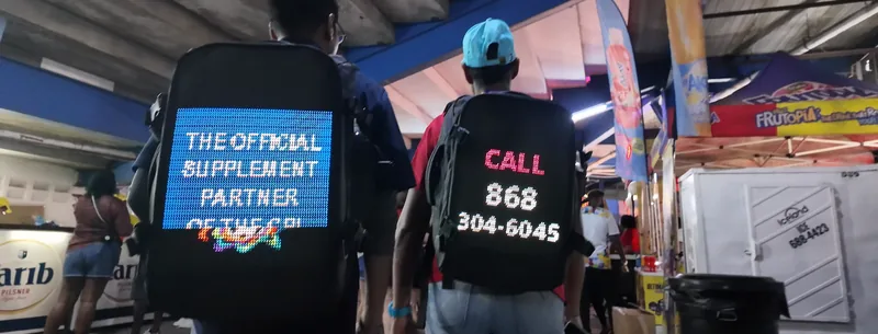 Two people walking a stadium concourse in Timesquare LED backpacks, one reading The Official Supplement Partner, the other Call 868 304-6045