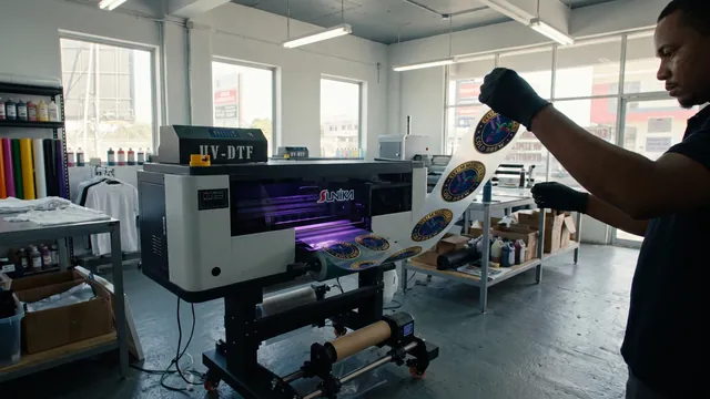 The UV DTF printer running under its violet curing lamp while an operator holds up a sheet of printed emblems
