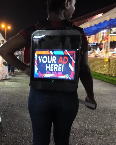 A person walking past market stalls at night wearing a lit LCD backpack reading Your Ad Here