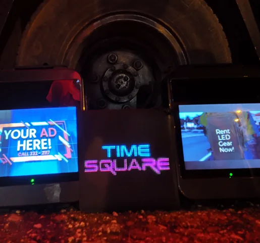 Two LCD backpacks and an LED sign panel reading Time Square, lit and standing on gravel at night, one backpack screen reading Your Ad Here, the other Rent LED Gear Now